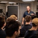 A briefing or meeting is taking place in what appears to be a military vessel's interior compartment. Two uniformed personnel, likely U.S. Navy sailors based on their dark blue coveralls and insignia patches, are standing and addressing a seated group of individuals. The setting has the characteristic appearance of a shipboard briefing room with metal walls, lockers, and a large display screen.