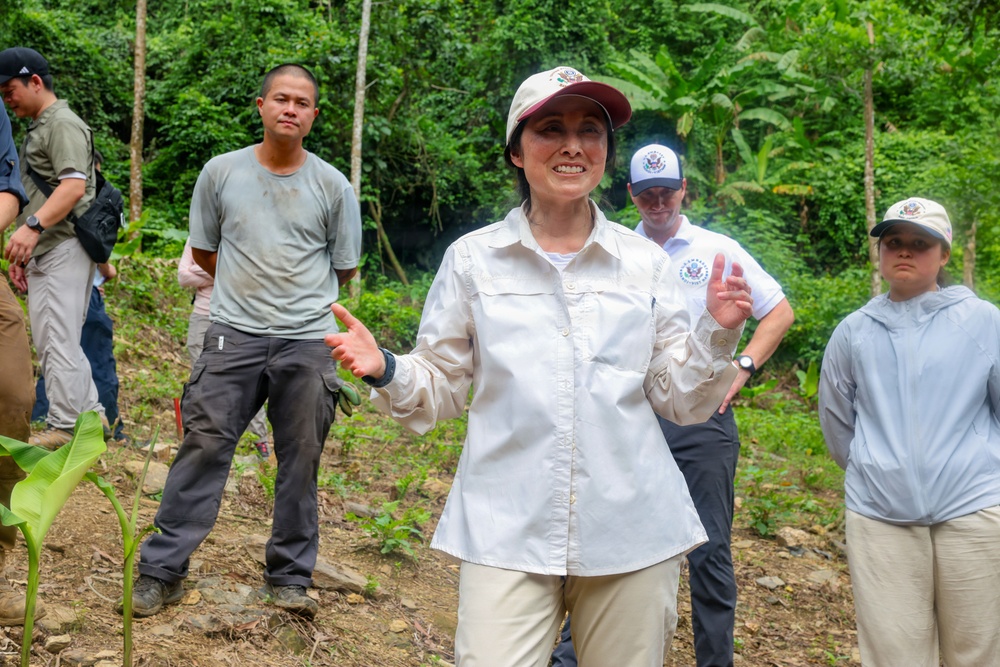 A group of people are gathered outdoors in a tropical forested area, likely on a field visit or official delegation. A woman in the foreground wearing a white field shirt and a cap with a dark brim appears to be speaking or presenting to the group. Several individuals in the background are wearing caps bearing what appears to be a U.S. government seal or emblem.
