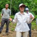 A group of people are gathered outdoors in a tropical forested area, likely on a field visit or official delegation. A woman in the foreground wearing a white field shirt and a cap with a dark brim appears to be speaking or presenting to the group. Several individuals in the background are wearing caps bearing what appears to be a U.S. government seal or emblem.