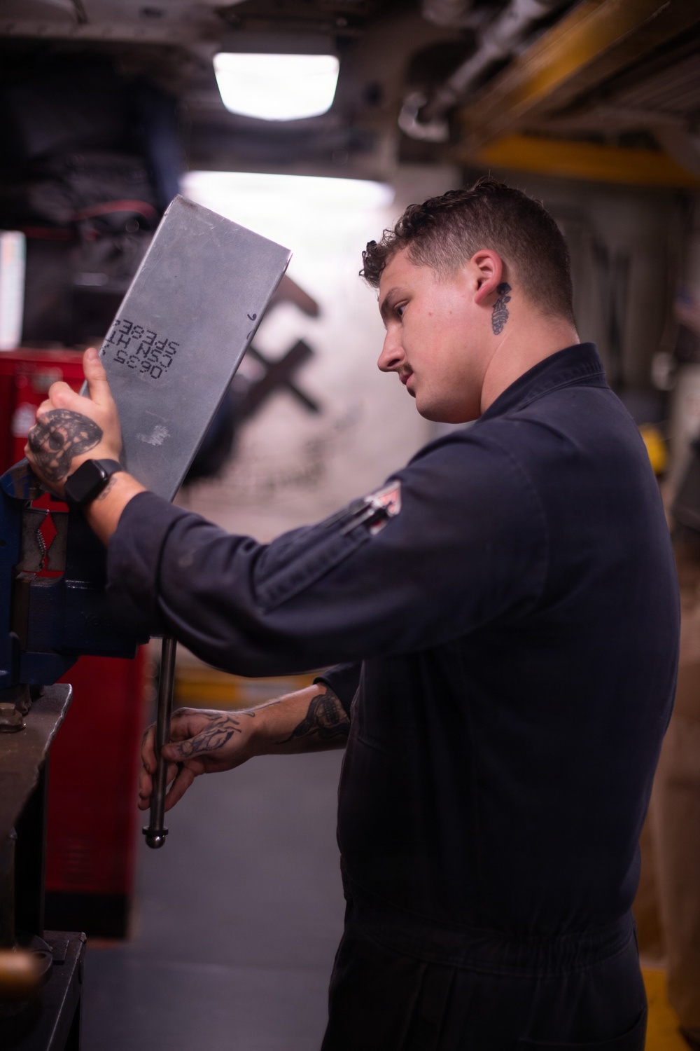 A young male mechanic or technician in a dark work uniform is handling a gray metal panel or access cover in what appears to be an automotive repair shop or industrial workspace. He has visible tattoos on both hands and forearms, a smartwatch on his left wrist, and an ear gauge. The metal panel has stamped or printed text/numbers on its surface.