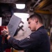 A young male mechanic or technician in a dark work uniform is handling a gray metal panel or access cover in what appears to be an automotive repair shop or industrial workspace. He has visible tattoos on both hands and forearms, a smartwatch on his left wrist, and an ear gauge. The metal panel has stamped or printed text/numbers on its surface.