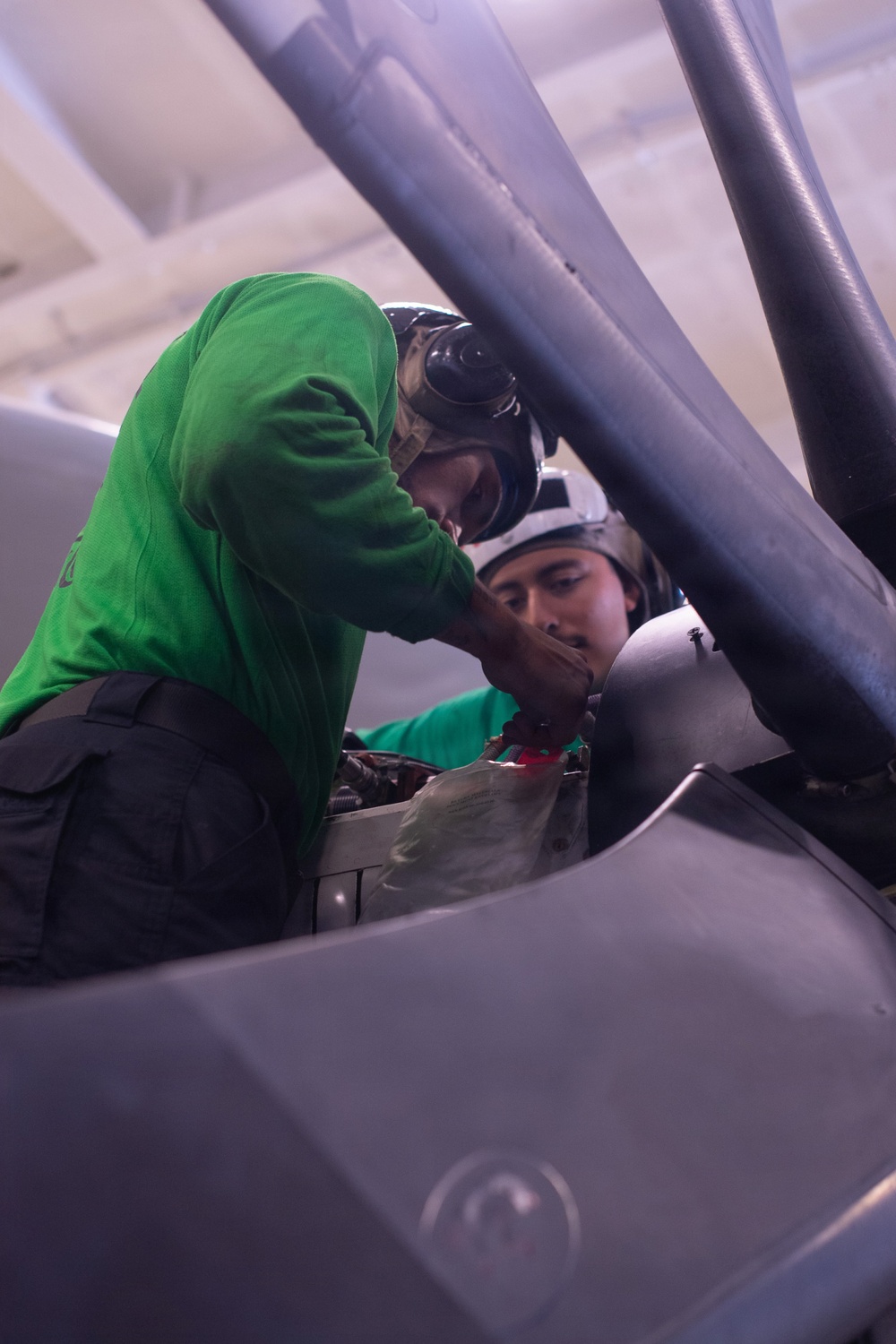 Two personnel wearing green jerseys and protective helmets are working inside or around the cockpit of a military aircraft. They appear to be performing maintenance or pre-flight checks on cockpit equipment. The image is taken from a close, angled perspective through the open canopy frame of the aircraft.