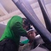 Two personnel wearing green jerseys and protective helmets are working inside or around the cockpit of a military aircraft. They appear to be performing maintenance or pre-flight checks on cockpit equipment. The image is taken from a close, angled perspective through the open canopy frame of the aircraft.