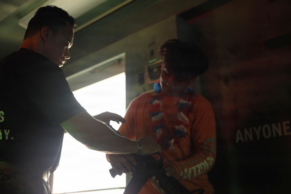 Two individuals are interacting in a dimly lit indoor space, with one person in a black shirt facing another wearing an orange shirt with a colorful lei or scarf. The scene appears to involve a hand-off or exchange of an object between the two people. A partially visible banner or poster with the word 'ANYONE' is visible on the right side of the frame.