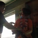 Two individuals are interacting in a dimly lit indoor space, with one person in a black shirt facing another wearing an orange shirt with a colorful lei or scarf. The scene appears to involve a hand-off or exchange of an object between the two people. A partially visible banner or poster with the word 'ANYONE' is visible on the right side of the frame.