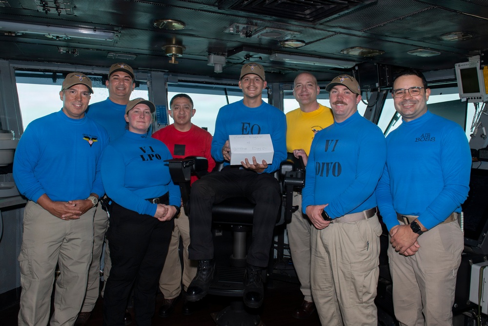 A group of approximately eight military personnel pose for a group photo on what appears to be the bridge of a naval vessel. Most are wearing blue long-sleeve shirts with role designations printed on them, along with tan/khaki pants and tan caps. One individual seated in the center holds a white box, suggesting a recognition or award ceremony.