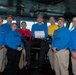 A group of approximately eight military personnel pose for a group photo on what appears to be the bridge of a naval vessel. Most are wearing blue long-sleeve shirts with role designations printed on them, along with tan/khaki pants and tan caps. One individual seated in the center holds a white box, suggesting a recognition or award ceremony.