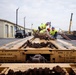 A group of workers in high-visibility safety vests are working on top of a flatbed railcar, handling heavy chains and securing equipment. The scene is set in an industrial yard with warehouse buildings, utility poles, and crane equipment visible in the background. The railcar has stenciled specifications including 'E69CE CPLR', '36 IN WHEELS', and 'NO. 24 BRAKE BEAMS' on its end panel.