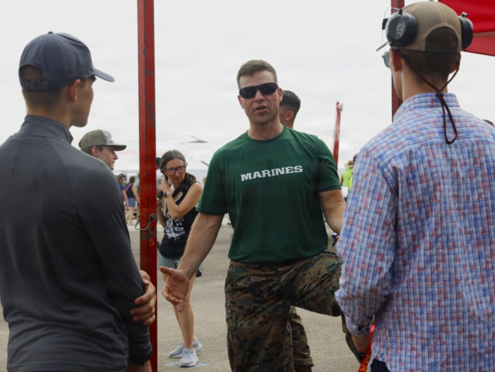 A U.S. Marine in a green 'MARINES' t-shirt and camouflage trousers is engaged in conversation with two civilians at what appears to be an outdoor public event or air show. The Marine appears to be in a recruiting or public outreach role, speaking with attendees near a red metal structure. The setting suggests a military open house or community engagement event.