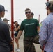 A U.S. Marine in a green 'MARINES' t-shirt and camouflage trousers is engaged in conversation with two civilians at what appears to be an outdoor public event or air show. The Marine appears to be in a recruiting or public outreach role, speaking with attendees near a red metal structure. The setting suggests a military open house or community engagement event.