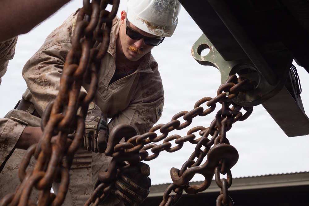 A military service member wearing desert camouflage uniform, white hard hat, and tactical gloves is working with heavy rust-covered chains attached to a large piece of equipment or vehicle. The low-angle shot emphasizes the chains in the foreground while the worker is focused on securing or rigging the chains to a metal bracket or hook. The setting appears to be an outdoor work area with a corrugated metal structure visible in the background.