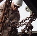 A military service member wearing desert camouflage uniform, white hard hat, and tactical gloves is working with heavy rust-covered chains attached to a large piece of equipment or vehicle. The low-angle shot emphasizes the chains in the foreground while the worker is focused on securing or rigging the chains to a metal bracket or hook. The setting appears to be an outdoor work area with a corrugated metal structure visible in the background.