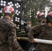 Military personnel in camouflage uniforms and red safety helmets are working around what appears to be a rocket artillery system, likely an M270 MLRS or similar multiple launch rocket system mounted on a vehicle. The personnel appear to be conducting loading, maintenance, or operational procedures with the launcher. A fire extinguisher is visible nearby as a safety precaution.