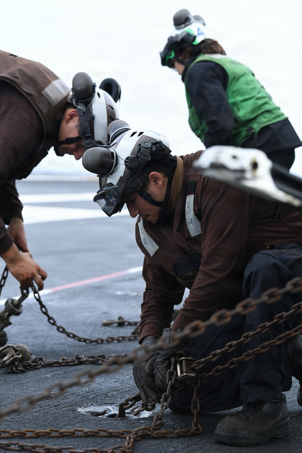 The image shows naval aviation deck crew members working on what appears to be an aircraft carrier flight deck. Personnel wearing brown jerseys and white helmets with hearing protection are handling heavy chains, likely tie-down or arresting gear chains. A third crew member in a green vest is visible in the background, indicating a supervisory or different functional role.