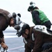 The image shows naval aviation deck crew members working on what appears to be an aircraft carrier flight deck. Personnel wearing brown jerseys and white helmets with hearing protection are handling heavy chains, likely tie-down or arresting gear chains. A third crew member in a green vest is visible in the background, indicating a supervisory or different functional role.