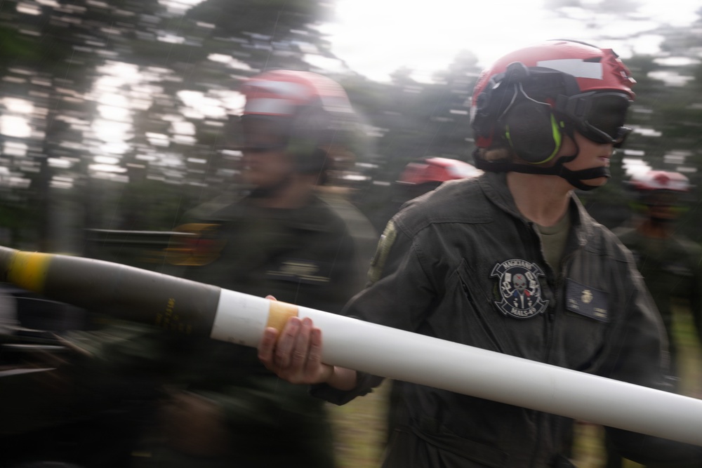Military personnel wearing red helmets and dark flight suits are carrying what appears to be a munition or ordnance component. The image is captured with motion blur, suggesting rapid movement during what appears to be an ordnance handling or loading operation. A unit patch is visible on the foreground figure's sleeve, consistent with a Marine aviation squadron.