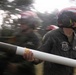 Military personnel wearing red helmets and dark flight suits are carrying what appears to be a munition or ordnance component. The image is captured with motion blur, suggesting rapid movement during what appears to be an ordnance handling or loading operation. A unit patch is visible on the foreground figure's sleeve, consistent with a Marine aviation squadron.