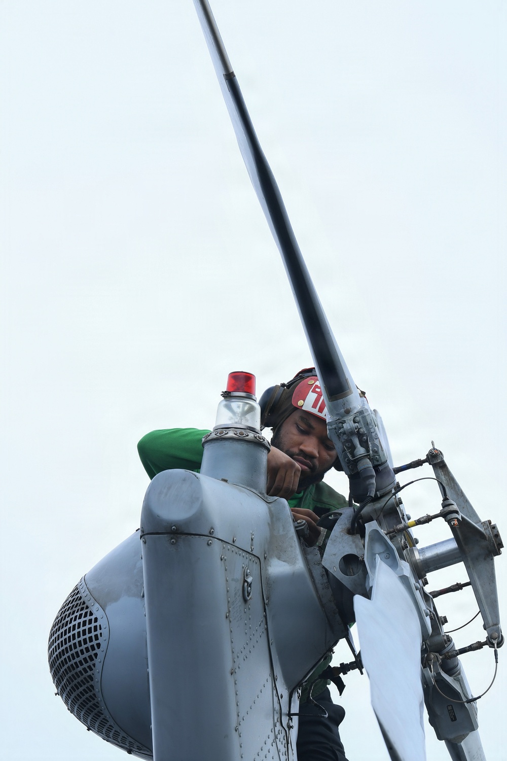 A technician wearing a green shirt and red helmet is performing maintenance or inspection work on what appears to be a helicopter rotor head assembly, viewed from a low angle looking upward. The worker is leaning over the top of the rotor hub, closely examining or working on mechanical components. A tall rotor mast extends upward into an overcast sky.
