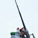 A technician wearing a green shirt and red helmet is performing maintenance or inspection work on what appears to be a helicopter rotor head assembly, viewed from a low angle looking upward. The worker is leaning over the top of the rotor hub, closely examining or working on mechanical components. A tall rotor mast extends upward into an overcast sky.