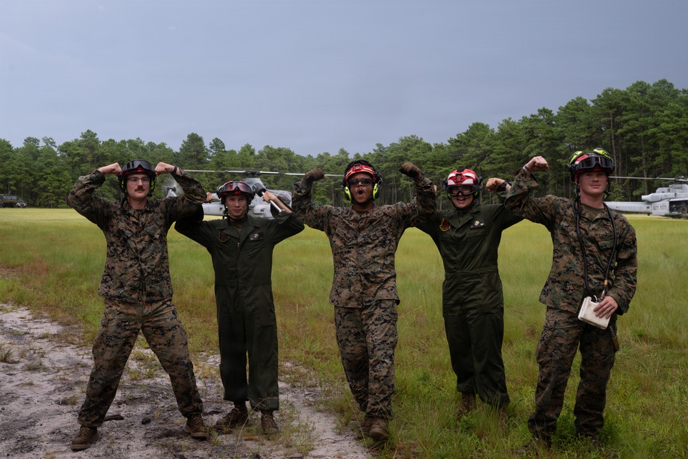 Five U.S. Marines pose together on a grassy field, flexing their arms in a celebratory manner. They are wearing a mix of MARPAT camouflage uniforms and olive drab flight suits, along with colored flight deck helmets with goggles. Military helicopters are visible in the background, suggesting a forward operating or training airfield.