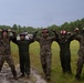 Five U.S. Marines pose together on a grassy field, flexing their arms in a celebratory manner. They are wearing a mix of MARPAT camouflage uniforms and olive drab flight suits, along with colored flight deck helmets with goggles. Military helicopters are visible in the background, suggesting a forward operating or training airfield.