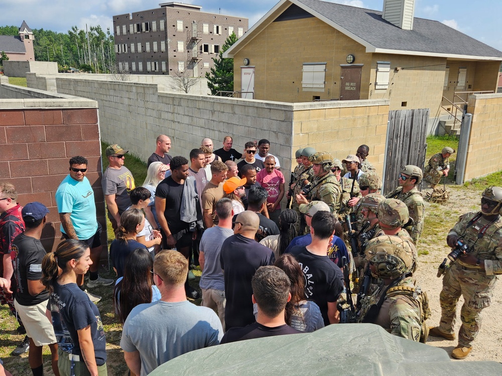 A group of civilians in casual clothing is gathered around several soldiers in full combat gear and camouflage uniforms at what appears to be a military training facility or mock urban environment. The soldiers are equipped with rifles, tactical vests, helmets, and communication gear, suggesting a demonstration or briefing is taking place. The setting includes concrete block walls, training buildings, and a controlled outdoor area consistent with a military base open house or public outreach event.