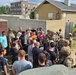 A group of civilians in casual clothing is gathered around several soldiers in full combat gear and camouflage uniforms at what appears to be a military training facility or mock urban environment. The soldiers are equipped with rifles, tactical vests, helmets, and communication gear, suggesting a demonstration or briefing is taking place. The setting includes concrete block walls, training buildings, and a controlled outdoor area consistent with a military base open house or public outreach event.
