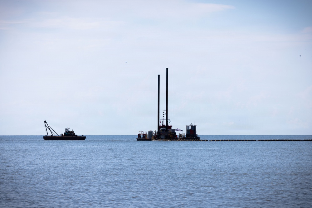 The image shows two industrial marine vessels operating on a calm, open body of water under an overcast sky. A large dredging platform with two tall vertical stacks dominates the center, while a smaller crane barge is positioned to the left. A floating pipeline or boom extends horizontally to the right of the dredging platform.