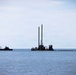 The image shows two industrial marine vessels operating on a calm, open body of water under an overcast sky. A large dredging platform with two tall vertical stacks dominates the center, while a smaller crane barge is positioned to the left. A floating pipeline or boom extends horizontally to the right of the dredging platform.