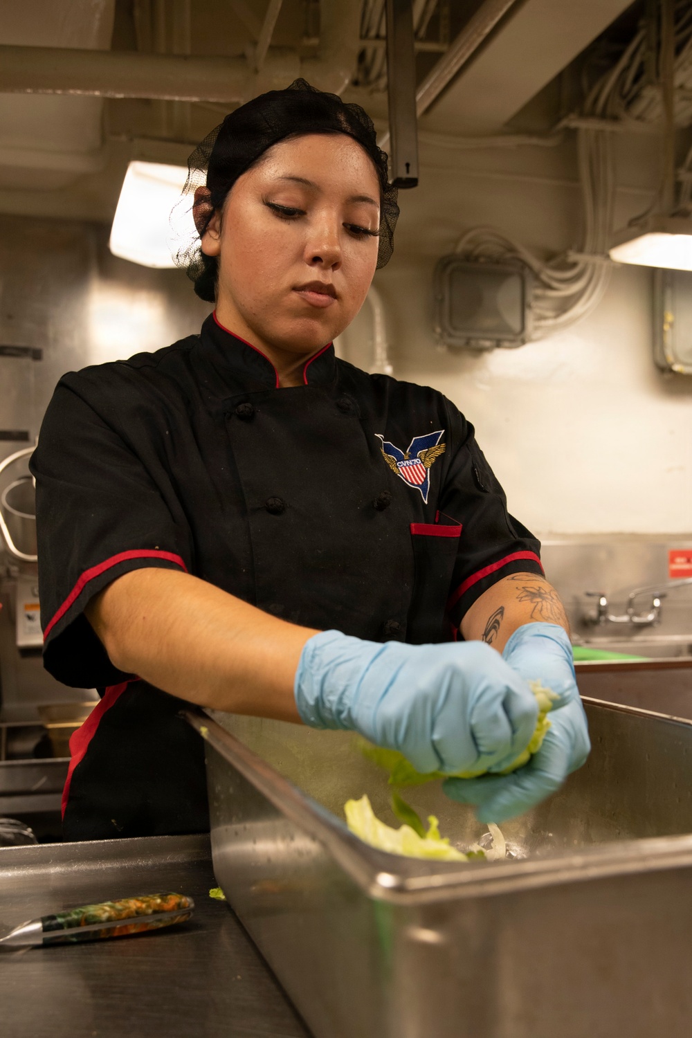 A female culinary specialist wearing a black chef's coat with a CVN-70 (USS Carl Vinson) emblem is preparing food in a military ship's galley kitchen. She is wearing blue nitrile gloves and a black hair net while handling what appears to be lettuce over a stainless steel food pan. The industrial kitchen environment and ship insignia indicate this is aboard a U.S. Navy aircraft carrier.
