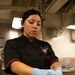 A female culinary specialist wearing a black chef's coat with a CVN-70 (USS Carl Vinson) emblem is preparing food in a military ship's galley kitchen. She is wearing blue nitrile gloves and a black hair net while handling what appears to be lettuce over a stainless steel food pan. The industrial kitchen environment and ship insignia indicate this is aboard a U.S. Navy aircraft carrier.