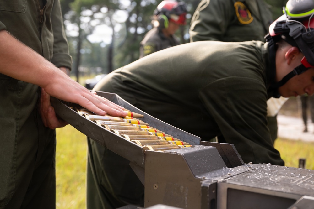 Military personnel in olive drab flight suits are handling a tray or magazine containing multiple rounds of ammunition with distinctive yellow and red band markings. The individuals are wearing hearing protection (ear defenders) positioned on their heads, suggesting proximity to loud equipment or weapons systems. The setting appears to be an outdoor military installation with trees visible in the background.