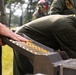 Military personnel in olive drab flight suits are handling a tray or magazine containing multiple rounds of ammunition with distinctive yellow and red band markings. The individuals are wearing hearing protection (ear defenders) positioned on their heads, suggesting proximity to loud equipment or weapons systems. The setting appears to be an outdoor military installation with trees visible in the background.