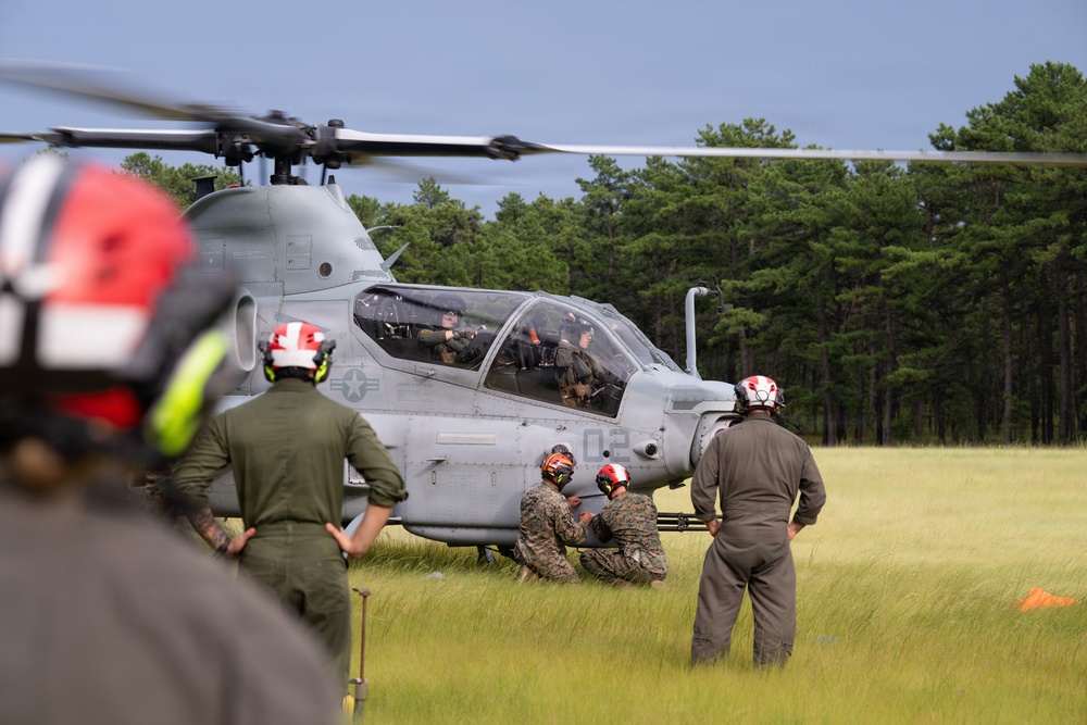 Military personnel in flight suits and helmets are gathered around a gray attack helicopter on a grassy field. The helicopter, marked with the number '02' and a U.S. Air Force star insignia, appears to be on the ground with its rotors spinning. Ground crew members wearing red and orange helmets are attending to the aircraft while two crew members are visible in the cockpit.