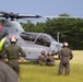 Military personnel in flight suits and helmets are gathered around a gray attack helicopter on a grassy field. The helicopter, marked with the number '02' and a U.S. Air Force star insignia, appears to be on the ground with its rotors spinning. Ground crew members wearing red and orange helmets are attending to the aircraft while two crew members are visible in the cockpit.