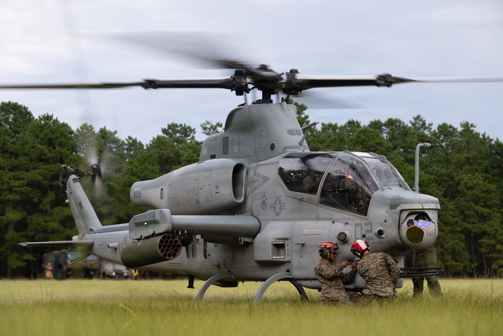 A U.S. Marine Corps AH-1Z Viper attack helicopter sits on a grassy field with its rotors spinning, indicating it is running or preparing for takeoff. Ground crew personnel in camouflage uniforms and flight helmets are crouched near the front of the aircraft, likely performing pre-flight checks or communications. The setting appears to be a forward operating area or training exercise location surrounded by pine trees.