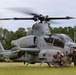 A U.S. Marine Corps AH-1Z Viper attack helicopter sits on a grassy field with its rotors spinning, indicating it is running or preparing for takeoff. Ground crew personnel in camouflage uniforms and flight helmets are crouched near the front of the aircraft, likely performing pre-flight checks or communications. The setting appears to be a forward operating area or training exercise location surrounded by pine trees.