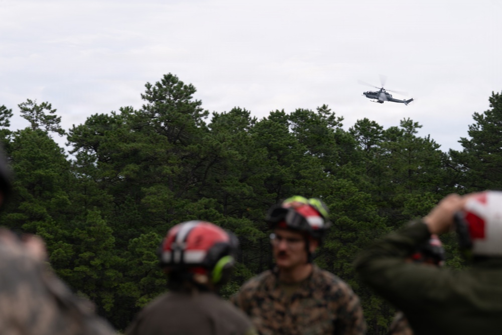 A group of military personnel wearing camouflage uniforms and red-accented helmets with hearing protection observe a helicopter flying overhead above a treeline. The helicopter appears to be a military attack or utility helicopter flying at low altitude. The setting appears to be a training or operational exercise in a forested area under overcast skies.