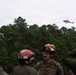 A group of military personnel wearing camouflage uniforms and red-accented helmets with hearing protection observe a helicopter flying overhead above a treeline. The helicopter appears to be a military attack or utility helicopter flying at low altitude. The setting appears to be a training or operational exercise in a forested area under overcast skies.