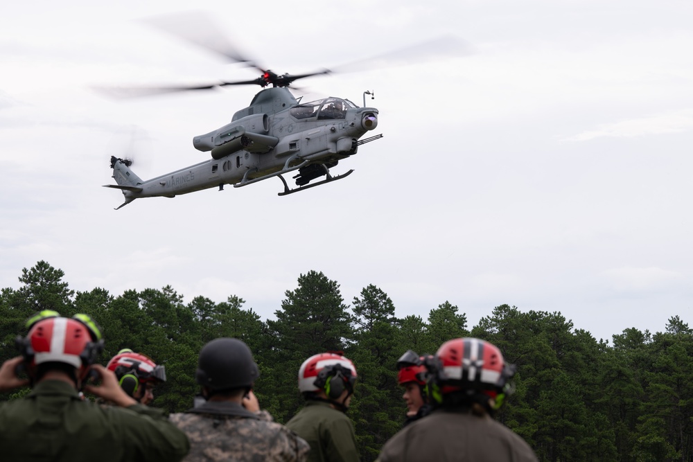 A U.S. Marines AH-1Z Viper attack helicopter is hovering at low altitude over a group of personnel wearing red flight deck helmets and military gear. The observers appear to be watching a demonstration or training exercise. The setting is an outdoor area with a treeline of pine trees in the background.