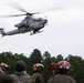 A U.S. Marines AH-1Z Viper attack helicopter is hovering at low altitude over a group of personnel wearing red flight deck helmets and military gear. The observers appear to be watching a demonstration or training exercise. The setting is an outdoor area with a treeline of pine trees in the background.