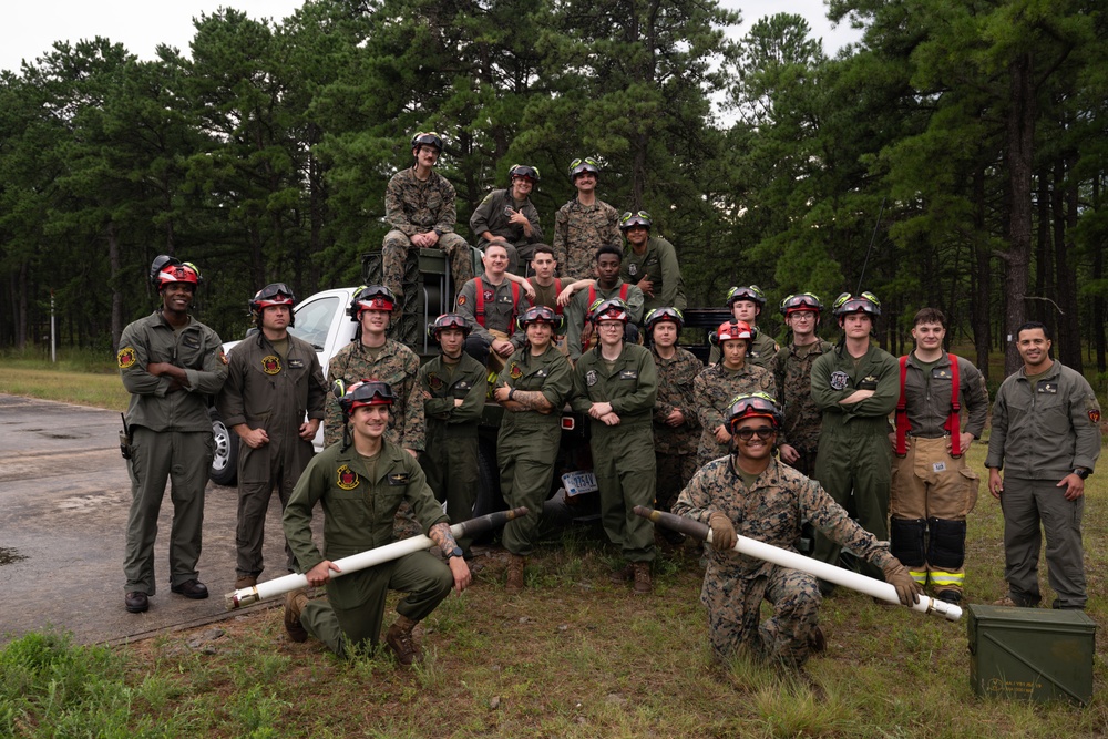 A group of approximately 20 military personnel, likely U.S. Marines, pose for a group photo outdoors near a military vehicle in a forested area. The personnel are wearing olive drab/green flight suits and coveralls, with several wearing red firefighting helmets with goggles. Two individuals in the foreground are kneeling and holding fire hose nozzles or similar equipment.