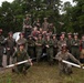 A group of approximately 20 military personnel, likely U.S. Marines, pose for a group photo outdoors near a military vehicle in a forested area. The personnel are wearing olive drab/green flight suits and coveralls, with several wearing red firefighting helmets with goggles. Two individuals in the foreground are kneeling and holding fire hose nozzles or similar equipment.