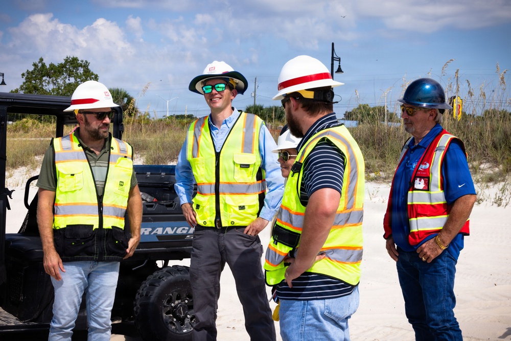 A group of five construction or site safety personnel are gathered outdoors near a sandy beach environment, engaged in conversation. They are wearing high-visibility safety vests and hard hats, suggesting an active worksite inspection or briefing. A Polaris Ranger utility vehicle is visible in the background.