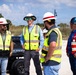 A group of five construction or site safety personnel are gathered outdoors near a sandy beach environment, engaged in conversation. They are wearing high-visibility safety vests and hard hats, suggesting an active worksite inspection or briefing. A Polaris Ranger utility vehicle is visible in the background.