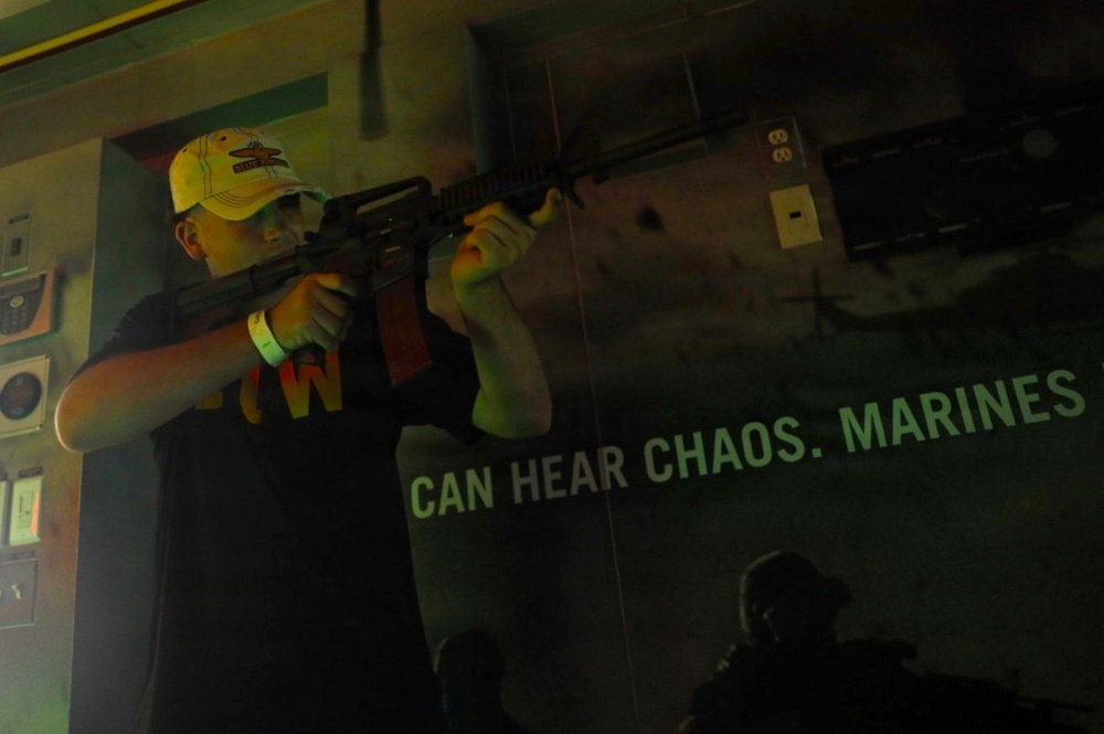 A person wearing a yellow baseball cap and dark t-shirt is holding and aiming a rifle-style firearm in a dimly lit indoor environment. Behind them, text on the wall reads 'CAN HEAR CHAOS. MARINES,' suggesting this is a military recruitment exhibit, simulator, or themed interactive display. The scene is lit with a greenish-yellow ambient light typical of experiential marketing or museum-style installations.