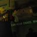 A person wearing a yellow baseball cap and dark t-shirt is holding and aiming a rifle-style firearm in a dimly lit indoor environment. Behind them, text on the wall reads 'CAN HEAR CHAOS. MARINES,' suggesting this is a military recruitment exhibit, simulator, or themed interactive display. The scene is lit with a greenish-yellow ambient light typical of experiential marketing or museum-style installations.