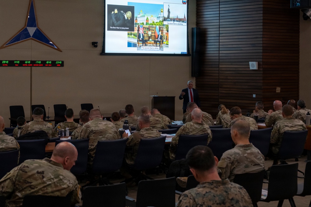 A civilian presenter in a suit delivers a briefing to a large audience of military personnel in camouflage uniforms in a formal conference room. The presentation slide on the large screen references images labeled 'The first Vlad,' 'The second Vlad,' and 'The third Vlad,' appearing to discuss Russian leadership or geopolitical topics. The room features a multi-timezone digital clock display on the left wall and a NATO-style emblem, suggesting an allied military headquarters environment.