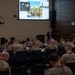 A civilian presenter in a suit delivers a briefing to a large audience of military personnel in camouflage uniforms in a formal conference room. The presentation slide on the large screen references images labeled 'The first Vlad,' 'The second Vlad,' and 'The third Vlad,' appearing to discuss Russian leadership or geopolitical topics. The room features a multi-timezone digital clock display on the left wall and a NATO-style emblem, suggesting an allied military headquarters environment.