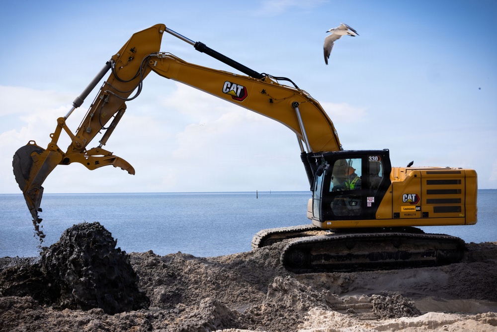 A yellow Caterpillar (CAT) 330 excavator is actively working on a coastal beach site, moving dark sediment or debris near the waterline. An operator is visible inside the cab, and a seagull is captured mid-flight above the machine. The background reveals a calm body of water under a partly cloudy blue sky.